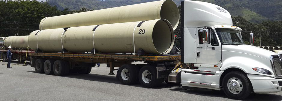 Camión blanco transportando grandes tuberías en una plataforma de carga en una carretera.