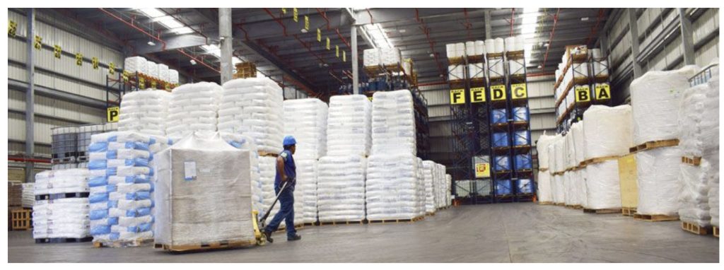 Interior de un almacén con palés apilados de productos, un trabajador con casco azul camina junto a ellos, señalización al fondo.