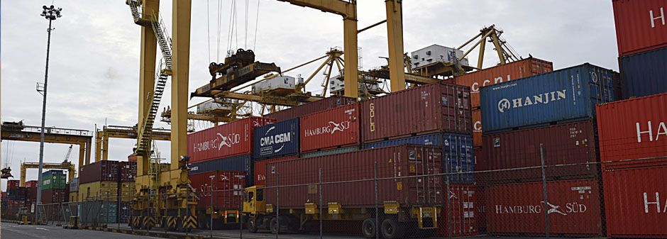 Contenedores apilados en un puerto con grúas grandes operando en el fondo.