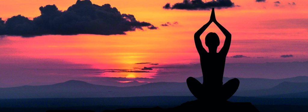 Persona en postura de meditación al atardecer con un cielo anaranjado y nubes oscuras.