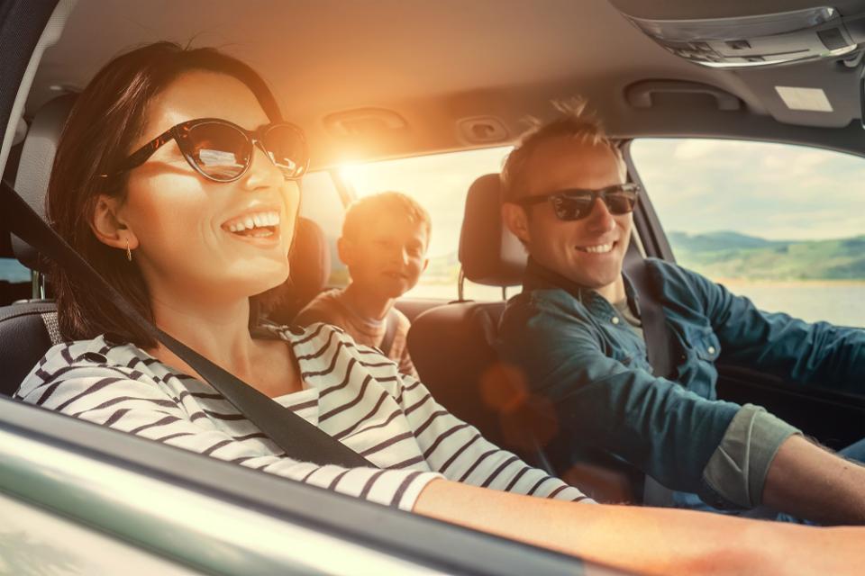Familia sonriente en un coche, el conductor y pasajera adelante con gafas de sol, un niño sentado atrás.