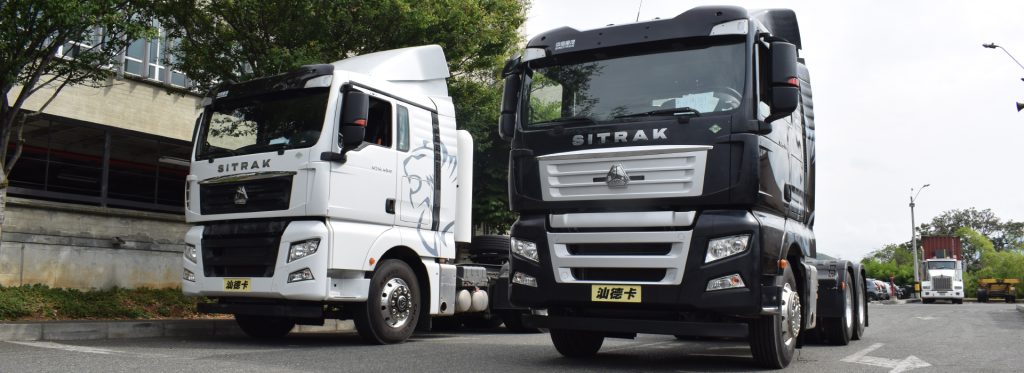 Dos camiones Sitrak, uno blanco y otro negro, estacionados en una calle con vegetación al fondo.