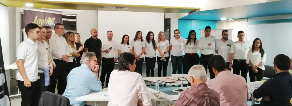 Grupo de personas en una presentación, la mayoría vistiendo camisas blancas, frente a una mesa con espectadores sentados.
