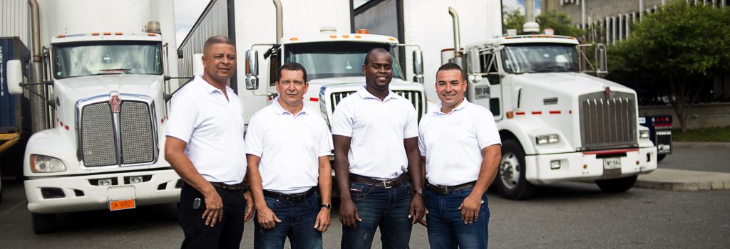 Cuatro hombres con camisetas blancas posan frente a camiones blancos estacionados.