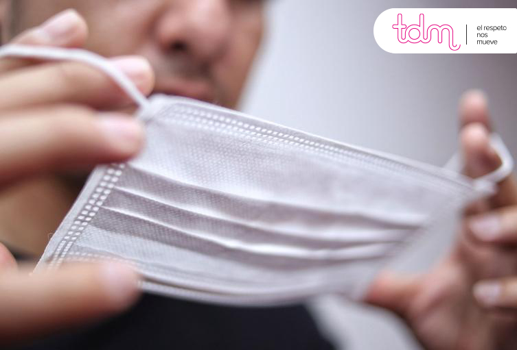 Persona sosteniendo una mascarilla quirúrgica blanca con un logotipo en la esquina superior derecha.