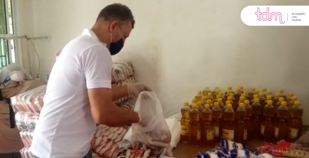 Persona con mascarilla y guantes empacando productos como aceite y otros alimentos en una bolsa.