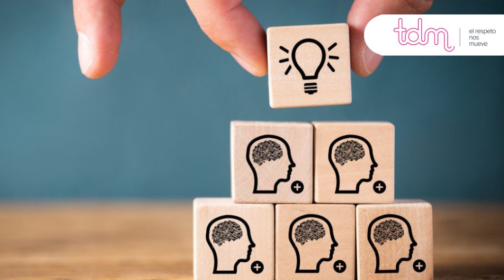 Una mano coloca un bloque de madera con una bombilla sobre otros bloques con cerebros, junto al logo de "tdm".