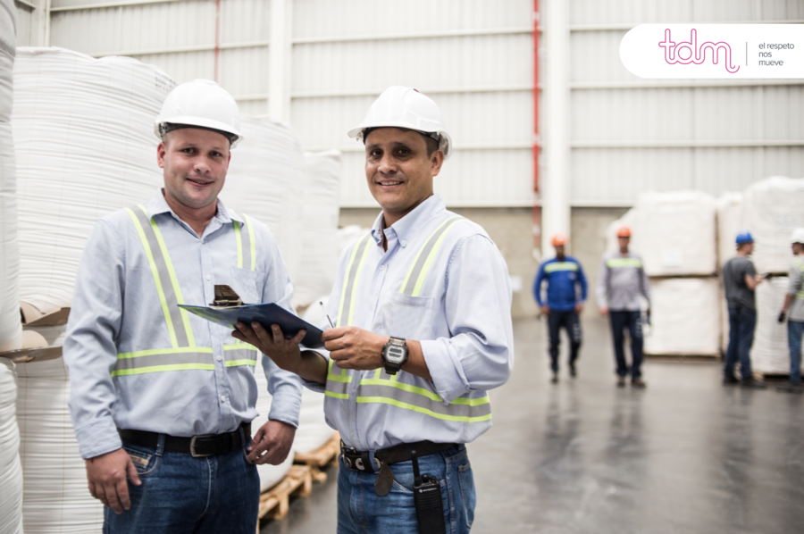 Dos hombres con cascos y chalecos reflectantes en un almacén, uno sosteniendo una carpeta. Al fondo, trabajadores con cascos.