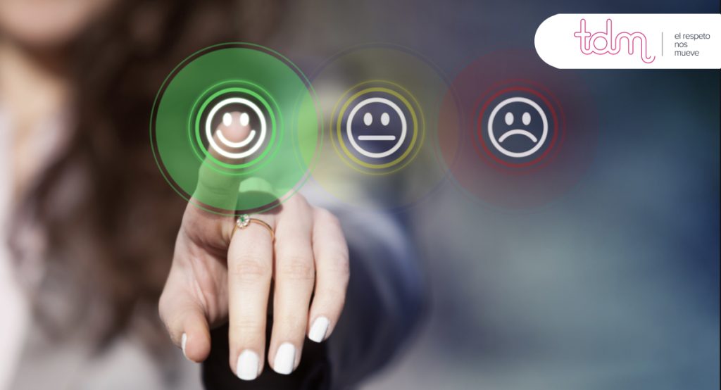 Mano apuntando a un emoticono sonriente iluminado en una pantalla, con otros dos emoticonos de expresión neutra y triste.