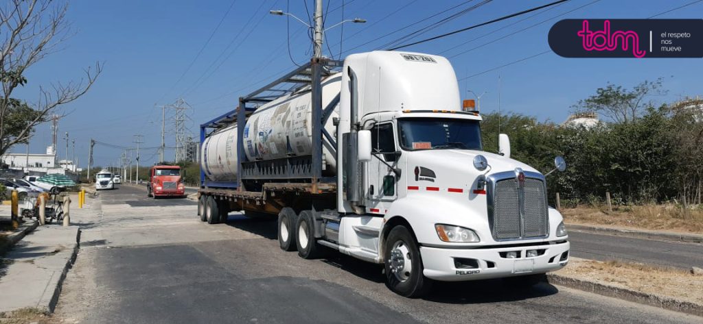 Camión blanco transportando un contenedor cilíndrico en un remolque, con otro camión rojo al fondo.