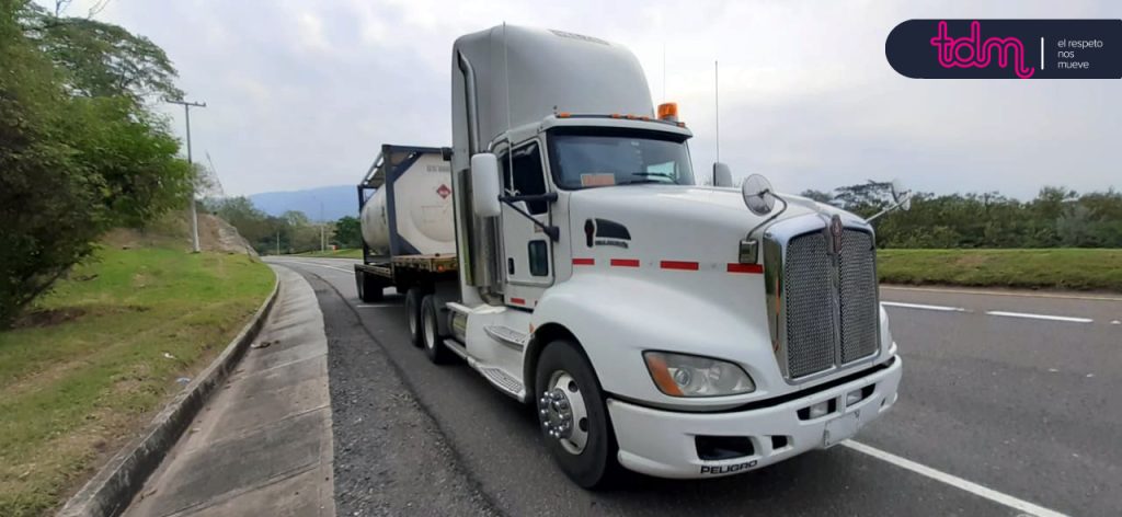 Camión blanco de carga con remolque en una carretera, rodeado de árboles y montañas al fondo.