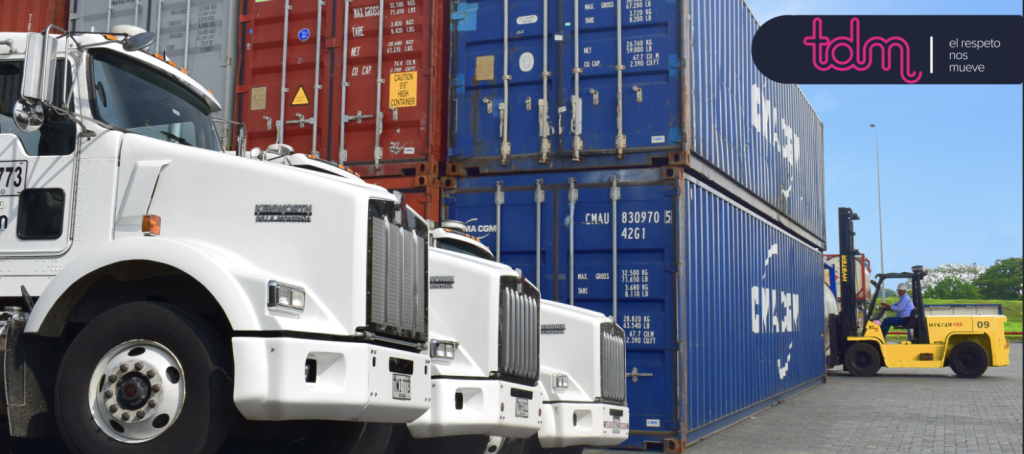 Camiones y montacargas en un puerto de contenedores con cajas de carga en rojo y azul; logo de tdm visible.