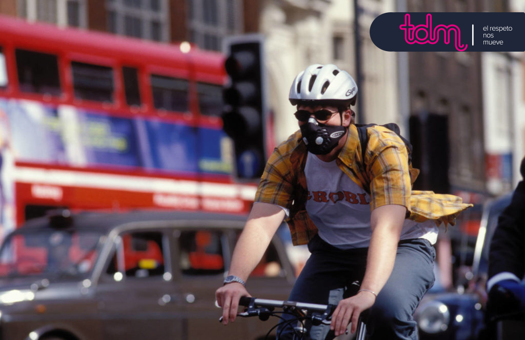 Persona en bicicleta con casco y mascarilla, en una calle urbana con un autobús rojo de dos pisos en el fondo.