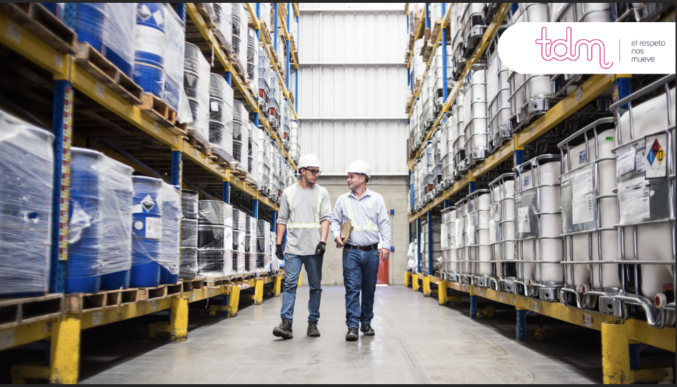 Dos hombres con cascos blancos caminan por un pasillo de un almacén, rodeados de grandes contenedores de líquido.
