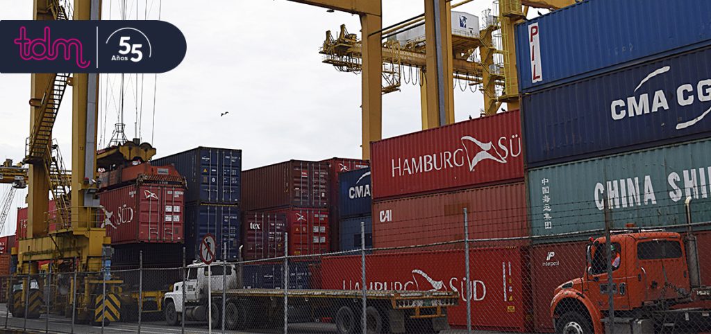 Contenedores apilados en un puerto con grúas amarillas y camiones cerca. Logos visibles de líneas navieras.