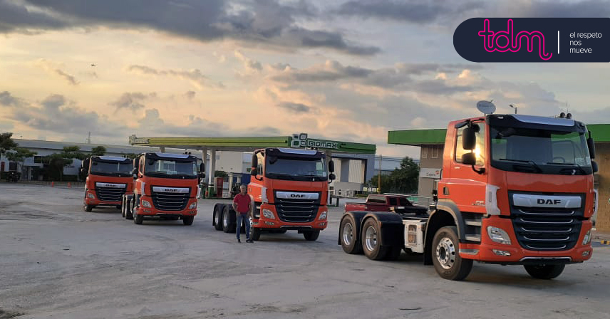 Cuatro camiones naranjas DAF en un estacionamiento cerca de una gasolinera al atardecer.