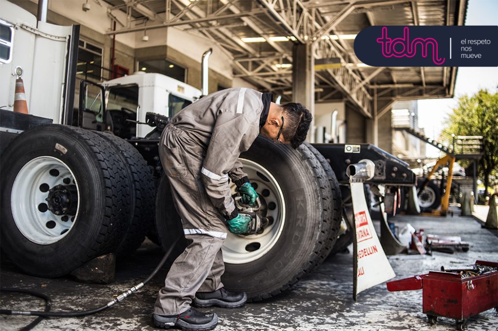 Un trabajador revisa un neumático grande en una estación de servicio, con herramientas y otros neumáticos alrededor.