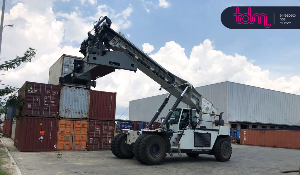 Grúa manipuladora elevando un contenedor en una zona de almacenamiento al aire libre, con contenedores apilados alrededor.