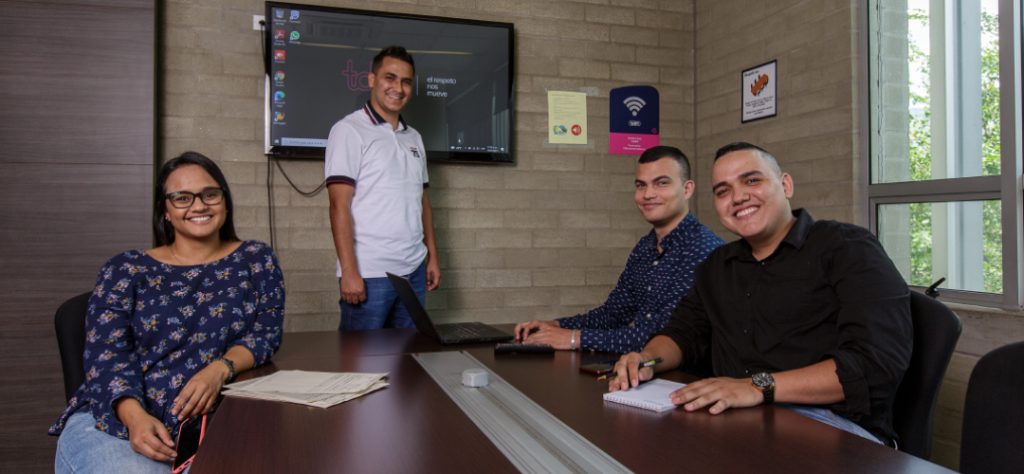 Cuatro personas sonríen en una sala de reuniones, con una pantalla y documentos sobre la mesa.