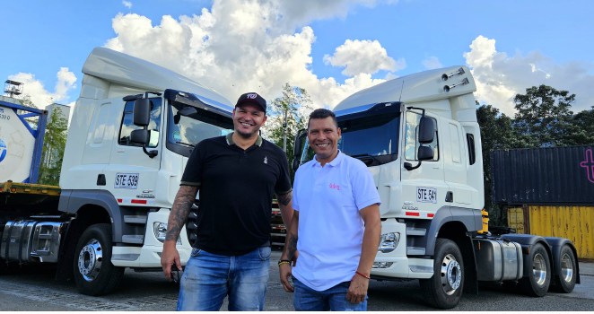 Dos hombres sonrientes de pie frente a dos camiones blancos estacionados con cielo azul de fondo.