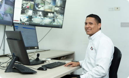 Hombre sonriendo en una oficina frente a una computadora, con pantallas de vigilancia en la pared.