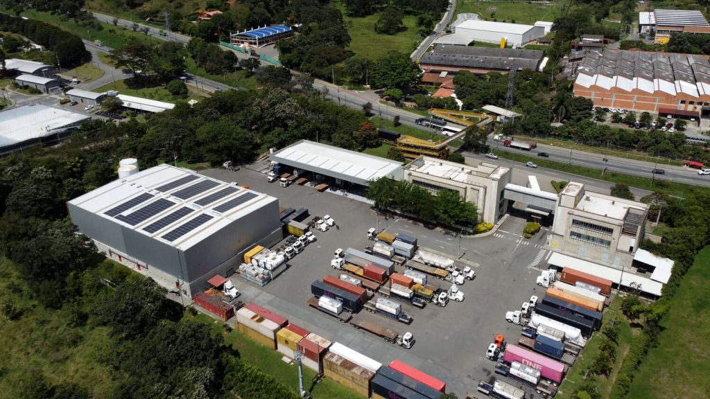 Vista aérea de un complejo industrial con varios edificios, contenedores de carga y camiones estacionados, rodeado de áreas verdes.