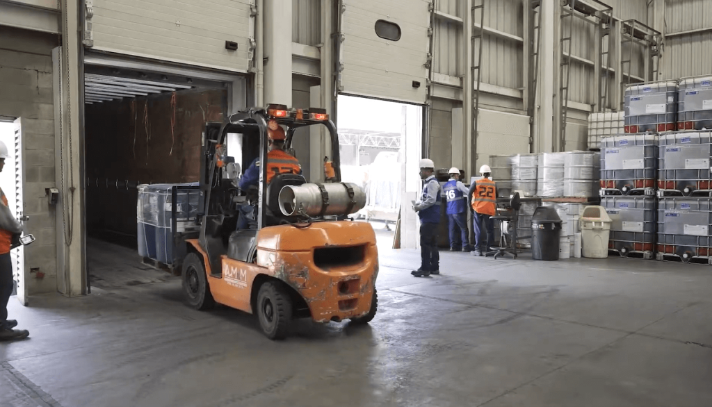 Una carretilla elevadora naranja cargando contenedores dentro de un almacén, mientras varios trabajadores observan.