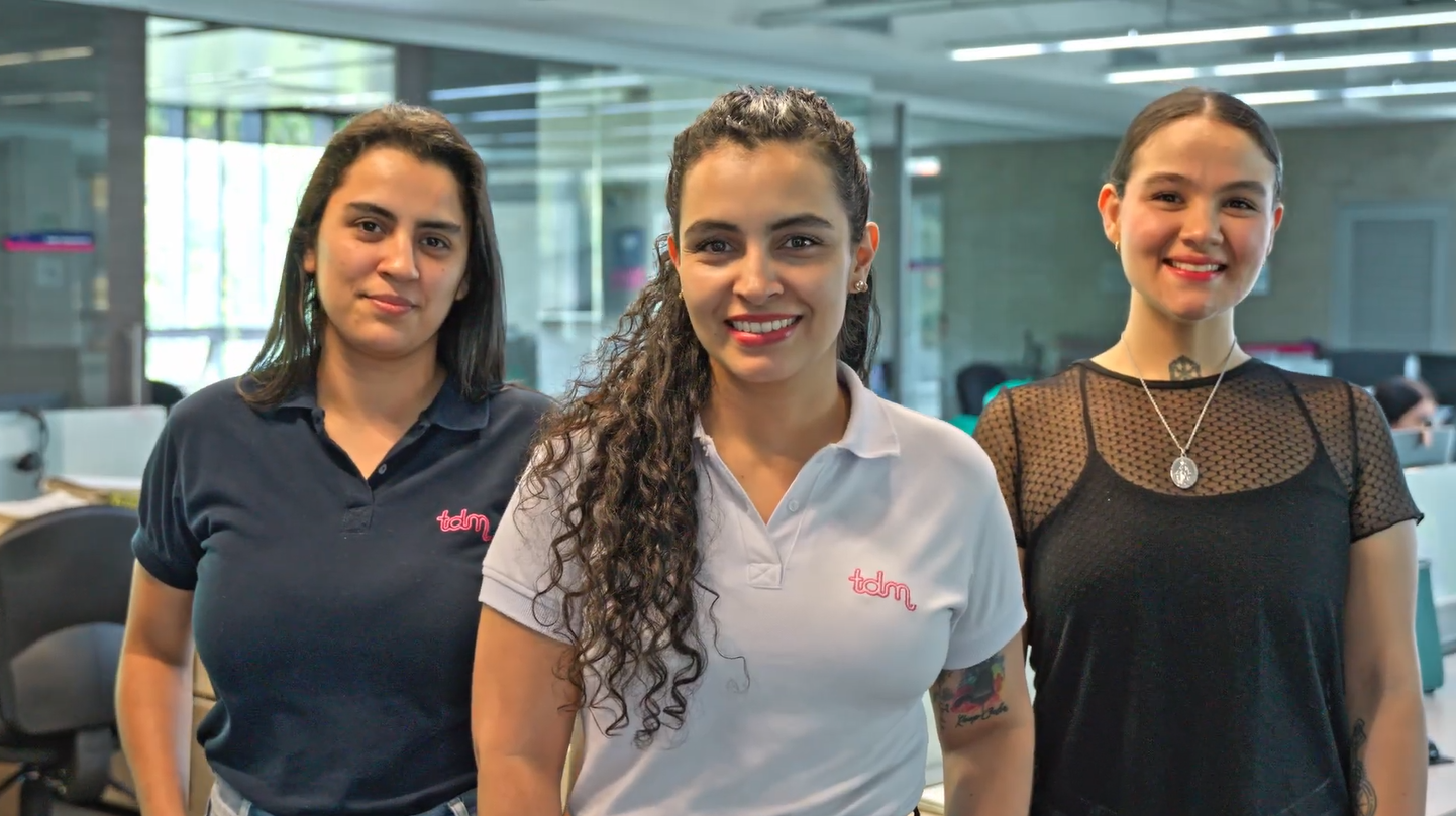 Tres mujeres posan sonrientes en una oficina moderna, usando camisetas corporativas.