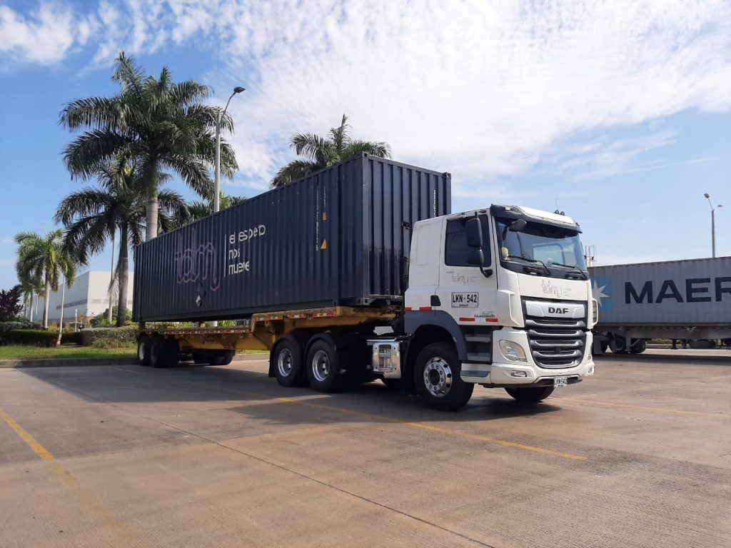 Camión de carga con contenedor azul oscuro estacionado en una zona pavimentada, rodeado de palmeras bajo un cielo parcialmente nublado.