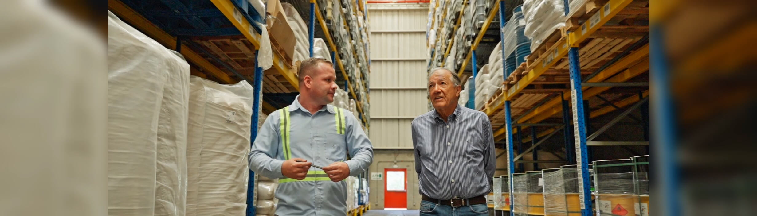 Dos hombres caminan y conversan en un almacén lleno de estantes altos con productos almacenados.