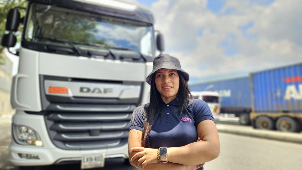 Mujer de pie frente a un camión blanco, con los brazos cruzados y sombrero. Otro camión se ve al fondo en un día soleado.