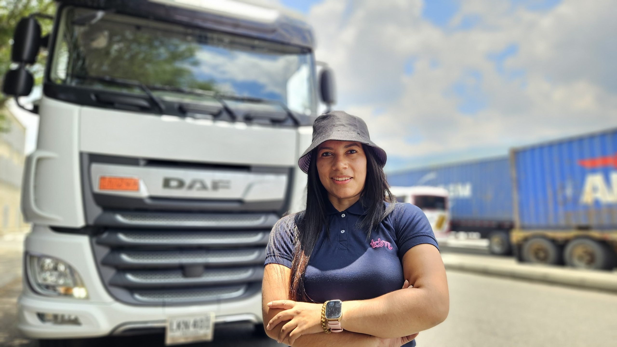 Mujer de pie frente a un camión blanco, con los brazos cruzados y sombrero. Otro camión se ve al fondo en un día soleado.